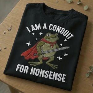 Peut inclure: Un t-shirt noir avec une broderie représentant une grenouille verte portant une cape rouge et tenant une épée argentée. Le texte "I AM A CONDUIT FOR NONSENSE" est brodé en blanc au-dessus et en dessous de la grenouille, entouré de petites étoiles.