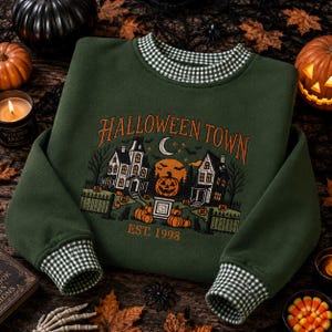 May include: A dark green sweatshirt with a black and white gingham collar and cuffs. The front features an embroidered scene of two houses, a crescent moon, and pumpkins with the text HALLOWEEN TOWN EST. 1998 in orange and white thread.