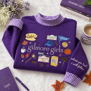 May include: A purple long-sleeve sweatshirt with a mock neck and cuffs featuring a purple and white gingham pattern. The front displays embroidered icons and the text "gilmore girls," while the sleeve features the text "where you lead i will follow."