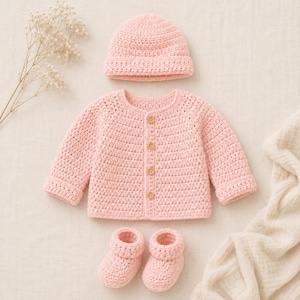 Pode incluir: Um conjunto de crochê rosa claro composto por um cardigã de manga comprida com quatro botões de madeira, um gorro a condizer e um par de botinhas. As peças apresentam um padrão de pontos texturizado consistente em todo o conjunto.