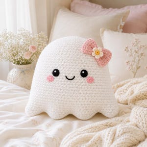 May include: A white crocheted ghost pillow with a rounded shape, featuring black embroidered eyes, a small black smile, and pink circular cheeks. A pink crocheted bow with a small white and yellow flower center is attached to the top right side.