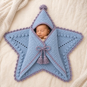 May include: A star-shaped crochet baby swaddle blanket in light blue with purple trim and a matching pom-pom at the top point. The blanket features a textured bobble stitch pattern, a scalloped edge, and a central tie closure with two tassels.