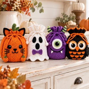 May include: Four crocheted drawstring bags featuring Halloween characters: an orange pumpkin cat, a white ghost with a purple bow tie, a purple one-eyed monster with green horns, and a black and orange owl. Each bag has a cinched top with drawstrings.