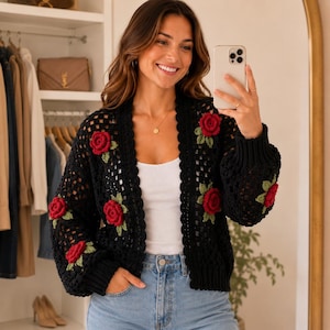 May include: A black crochet cardigan featuring a square-patterned knit design with three-dimensional red rose appliqués and green leaves. The garment has long sleeves with ribbed cuffs, an open front, and a scalloped edge along the neckline.