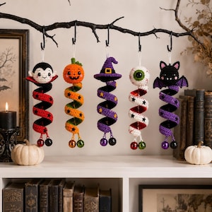 May include: Five hanging crochet ornaments featuring a vampire, a pumpkin, a witch hat, an eyeball, and a bat. Each figure is attached to a colorful, spiraling yarn tail decorated with small black stars and finished with two small metal jingle bells.