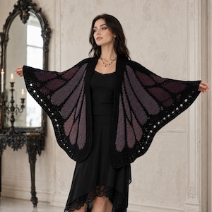 Puede incluir: Un chal de ganchillo en negro y morado oscuro diseñado para parecerse a alas de mariposa. La prenda presenta un borde negro sólido y un patrón texturizado en forma de ala en tonos morados y negros, usado sobre una blusa negra sin mangas.