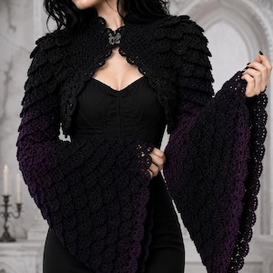 Op de afbeelding: Een zwart gehaakte bolero met een textuur van schubben en wijde, uitlopende mouwen die bij de boorden overgaan in een dieppaarse kleur. Het kledingstuk wordt bij de halslijn gesloten met een decoratieve zilverkleurige metalen sluiting.
