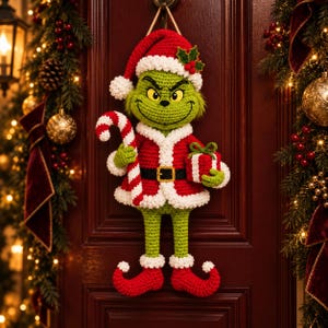 Könnte beinhalten: Ein gehäkelter Wandbehang in Form einer grünen Kreatur, die einen rot-weißen Weihnachtsmannanzug, eine passende Mütze mit Stechpalmenblatt und Stiefel mit gekräuselten Spitzen trägt. Die Figur hält eine rot-weiß gestreifte Zuckerstange und ein Geschenk.