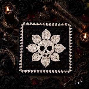 Może przedstawiać: Kwadratowy czarny szydełkowy kwadrat typu granny square z centralnym motywem białej czaszki otoczonym ośmioma białymi szydełkowanymi liśćmi. Kwadrat jest wykończony delikatną białą falistą krawędzią, tworząc kontrastowy wzór o wyraźnej fakturze.