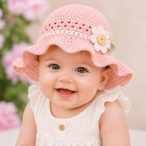 May include: A light pink crochet sun hat featuring a textured stitch pattern and a white daisy appliqué with a yellow center and a small green leaf. The hat has a wide, wavy brim and a decorative band around the crown, crafted from soft yarn.