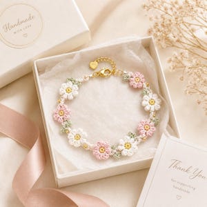 May include: A crochet flower bracelet featuring alternating white and light pink blossoms with yellow centers and small green leaves. The bracelet includes a gold-toned metal lobster clasp and a small heart-shaped charm, presented in a white gift box.