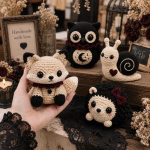 Puede incluir: Una colección de cuatro animales de peluche tejidos a ganchillo que incluye un zorro beige con cuello rojo oscuro, un búho negro con un parche de luna y estrella, un caracol beige con concha negra y corazón, y un erizo negro con lazo rojo.