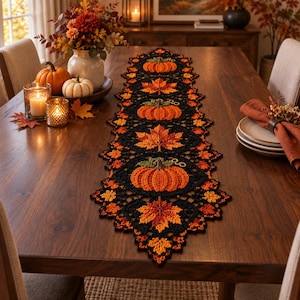 Puede incluir: Un camino de mesa de ganchillo largo y rectangular con un patrón repetitivo de calabazas naranjas y hojas de arce otoñales multicolores en tonos rojos, naranjas y amarillos, enmarcado por un borde de hilo negro con un acabado festoneado.
