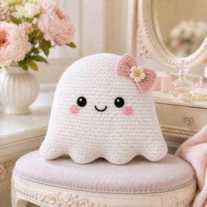 May include: A white crocheted ghost plush toy featuring a rounded shape with a scalloped bottom edge. The ghost has two black circular eyes, a small stitched smile, and two pink circular cheeks. A pink crocheted bow with a small white flower is attached.