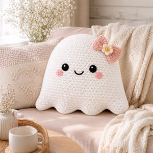 May include: A white crochet ghost plushie featuring black embroidered eyes, a small black smile, and pink circular cheeks. A light pink crocheted bow with a small white and yellow flower center is attached to the top right side of the ghost head.