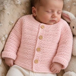May include: A light pink long-sleeve cardigan made of crocheted yarn. The garment features a round neckline and a front button closure with four small, round, light brown wooden buttons. The sleeves have folded cuffs at the wrists.