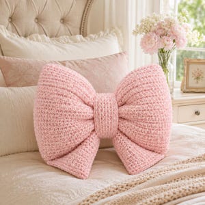 May include: A light pink, textured, crocheted throw cushion shaped like a large bow. The cushion features a dense, uniform knit pattern throughout its surface, creating a soft and dimensional appearance with two wide loops and a central knot detail.