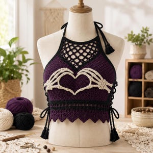 May include: A dark purple crochet halter top featuring a cream-coloured skeleton hand design forming a heart shape at the centre. The garment has a black lattice neckline, black trim, and side ties with tassels, displayed on a mannequin torso.