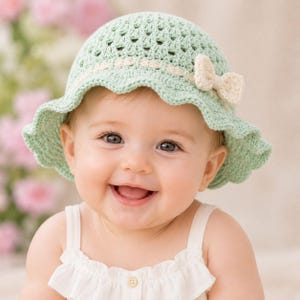 Puede incluir: Un sombrero de sol de ganchillo en color verde salvia claro con un patrón de punto calado, un ala festoneada y una cinta tejida de color crema decorada con un pequeño lazo de ganchillo a juego en el lateral del sombrero.