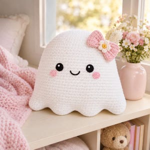 May include: A white crocheted ghost plush toy featuring a smiling face with black eyes, pink circular cheeks, and a pink bow accessory on the side adorned with a small white and yellow flower. The ghost has a rounded top and a scalloped bottom edge.