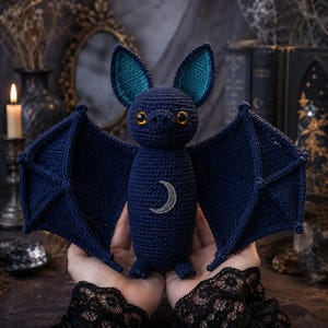 May include: A dark blue crochet bat plush toy featuring large pointed ears with teal inner lining, bright orange eyes, and a silver crescent moon emblem centered on its chest. The bat has wide, outstretched wings and small feet.