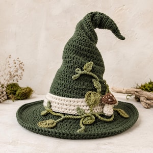 May include: A dark green crocheted pointed hat featuring a wide brim and a tall, slightly curved crown. A cream-colored band encircles the base, decorated with crocheted green vines, leaves, and a small brown mushroom with white spots.