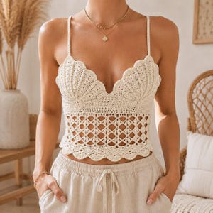 May include: A cream-colored crochet crop top featuring a shell-inspired bodice design with thin straps and a scalloped hemline. The midsection of the garment is constructed with an open diamond-shaped lattice pattern, providing a textured look.