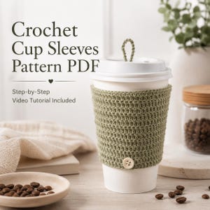May include: A white paper coffee cup with a green crocheted sleeve and a button closure. The text "Crochet Cup Sleeves Pattern PDF" is displayed above the cup. The image also includes a small plate with coffee beans.