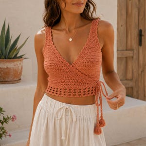 May include: A terracotta-colored crochet crop top featuring a V-neckline, a wrap-style front with side tie closures, and a scalloped hem. The garment is crafted with a textured stitch pattern and includes long, dangling tassels at the waist ties.