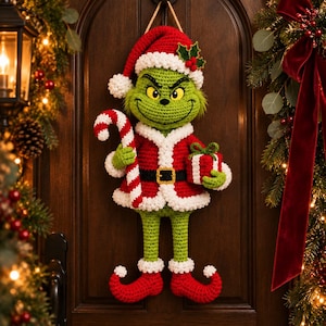 May include: A crocheted wall hanging of a green character wearing a red and white Santa suit, a matching hat with holly, and curled-toe boots. The figure holds a red and white striped candy cane in one hand and a small red gift box in the other.