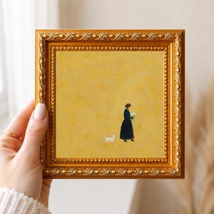 May include: A square painting in a decorative gold frame with a textured yellow background. The artwork depicts a person in a dark green dress holding white flowers, accompanied by a small white cat walking on the ground in the lower center.