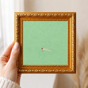 May include: A square art print featuring a small figure swimming in a solid light green field, encased in an ornate, metallic gold frame with a beaded inner border and a decorative, textured outer edge, presented in a square orientation.
