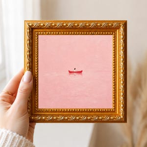 May include: A square painting in an ornate gold frame featuring a small red canoe with a single figure floating in the center of a vast, textured light pink expanse. The artwork displays a minimalist composition with a solid pastel pink background.