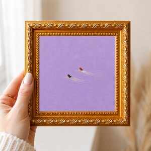 May include: A square painting in a decorative gold frame featuring two figures swimming in a solid lavender-colored expanse. The figures are depicted from above, with one wearing a light green swimsuit and the other wearing a white swimsuit.