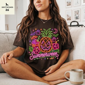 May include: A dark brown short-sleeve t-shirt featuring a neon-style graphic print. The design includes a jack-o'-lantern pineapple, tropical flowers, a cocktail, ghosts, bats, and stars. The text Summerween is written in a neon pink script.