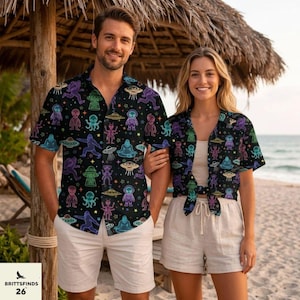May include: A black short-sleeved button-down shirt featuring a vibrant, neon-colored pattern of various aliens, UFOs, and Bigfoot figures scattered across the fabric. The design includes purple, green, blue, and yellow outlines on a dark background.