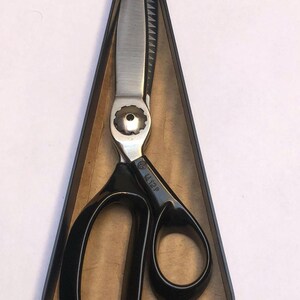 Könnte beinhalten: Professionelle Kleiderscheren mit silberner Klinge und schwarzen Griffen. Die Schere befindet sich in einem dreieckigen Etui. Die Klinge hat eine gezahnte Kante. Der Griff trägt die Aufschrift "44 12 P".