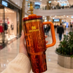 Könnte beinhalten: Ein durchscheinender orangefarbener Stanley-Becher mit Blumenmuster, einem massiven orangefarbenen Griff und einem passenden Deckel. Ein goldfarbener Metallaufsatz in Ananasform ist oben auf dem Deckel befestigt. Der Markenname Stanley ist in Gold gedruckt.