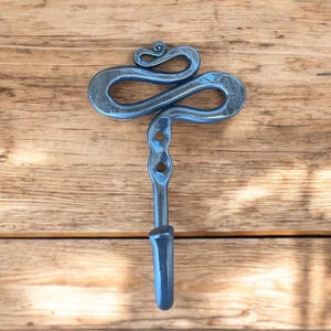 Puede incluir: Un gancho de metal azul con un diseño decorativo en espiral. El gancho está montado en una superficie de madera.