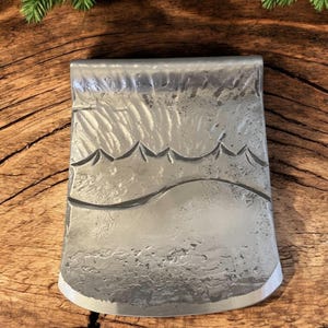 Op de afbeelding: Een zilveren bijlkop met een bergketenontwerp. De bijlkop ligt op een houten oppervlak.