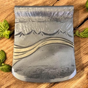 Op de afbeelding: Een zilveren metalen geldclip met een getextureerd oppervlak en een gouden ontwerp van bergen en een rivier.