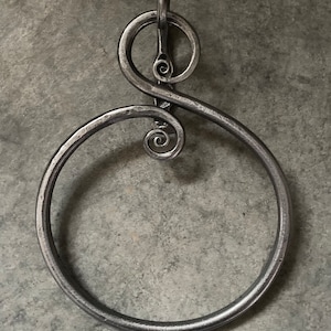 Pode incluir: Um porta-toalhas de metal prateado feito à mão com um design decorativo em espiral. O anel é circular com um laço menor na parte superior para pendurar. O metal tem uma aparência texturizada e martelada, sugerindo um estilo rústico e artesanal.