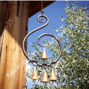 May include: A handcrafted metal wind chime with a unique spiral design. The chime features four bronze-coloured bells suspended from a decorative metal frame. The background includes a wooden structure, blue sky, and green foliage.