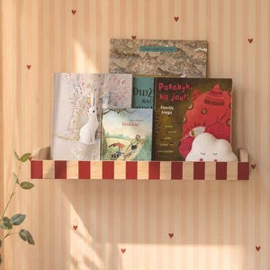 May include: A wooden shelf with a red and white striped front holds several children's books. A white rabbit figurine and a cloud-shaped night light are also on the shelf. The background features a striped wallpaper with small red hearts.
