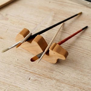 Handcrafted maple wood camel-shaped paintbrush holder on a light vintage wooden desk. The camel securely holds several art brushes and a pencil between its humps. The background features a cozy illustrator's workspace with a sketchpad.