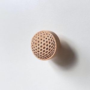 Peut inclure: Un bouton de tiroir rond en bois avec un motif texturé de balle de golf. Le bouton est brun clair et est monté sur un tiroir blanc.