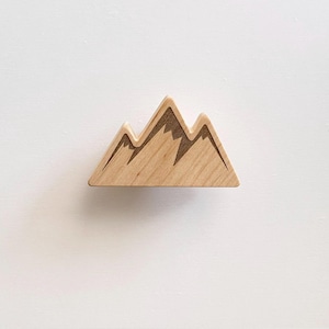 May include: A wooden mountain-shaped drawer pull with a natural wood finish and dark brown etched details. The drawer pull is mounted on a white cabinet, offering a rustic and outdoorsy aesthetic. The mountain design adds a touch of nature to the furniture.