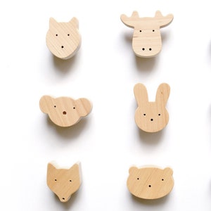 May include: Nine wooden animal-shaped knobs, including a cat, moose, monkey, koala, rabbit, giraffe, fox, bear, and sheep. Each knob is light brown with black dot eyes and a small hole for mounting. The knobs are arranged in a grid pattern on a white background.