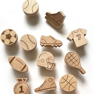 Peut inclure: Ornements en bois sur le thème du sport, comprenant un ballon de baseball, un ballon de football américain, un ballon de basket-ball, un ballon de football, une raquette de tennis, un patin à glace, un trophée et une médaille. Les ornements sont tous en bois clair et ont une finition naturelle.