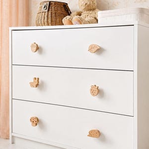 Op de afbeelding: Een witte ladekast met drie lades en zes lichtgekleurde houten knoppen in de vorm van een eikel, een blad, een konijn, een aardbei, een paddenstoel en een egel. De kast heeft een strak, rechthoekig frame met horizontale ladefronten.